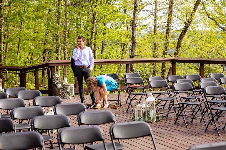 funny moment at wedding ceremony while man waits for his partner as she fixes her shoes, surrounded by empty chairs, with his suit coat thrown over his shoulder and an impatient look on his face.