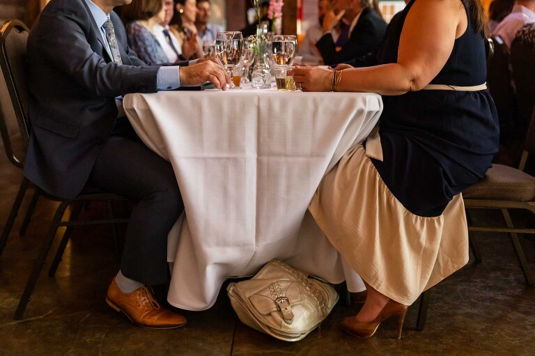 quiet moment at a table, with man and woman sitting across from each other and purse resting between them. up close detail of their bottom half captured by candid wedding photographer from pittsburgh, pamela anticole