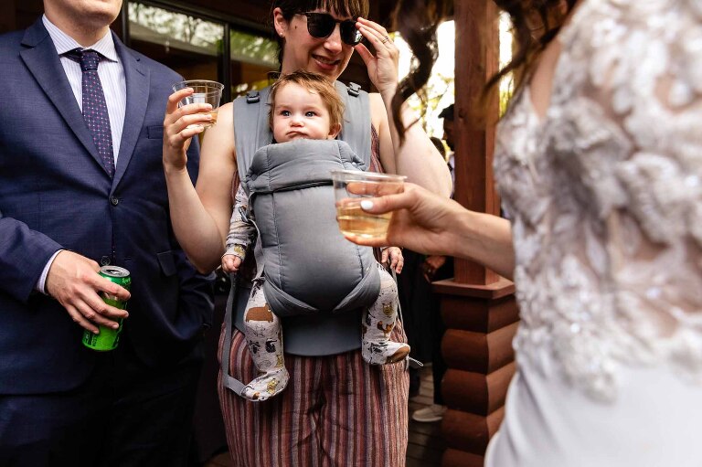 funny photo of baby in carrier outside on porch surrounded by adults holding alcohol at a wedding cocktail hour, photographed in a candid way by a photojournalist wedding photographer