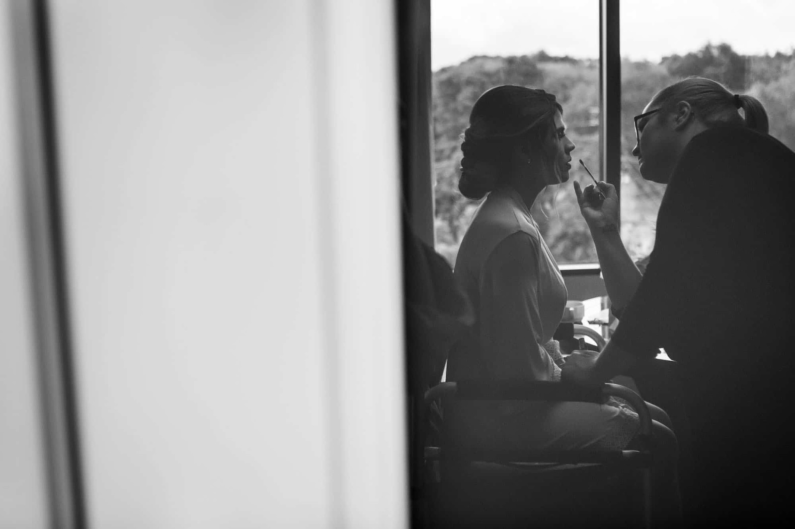 black and white photograph of bride getting her makeup done in front of a window.
