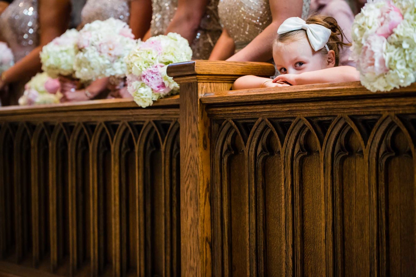 flower girl rests her head on her hands, half asleep during wedding ceremony while surrounded by bridesmaids at church.