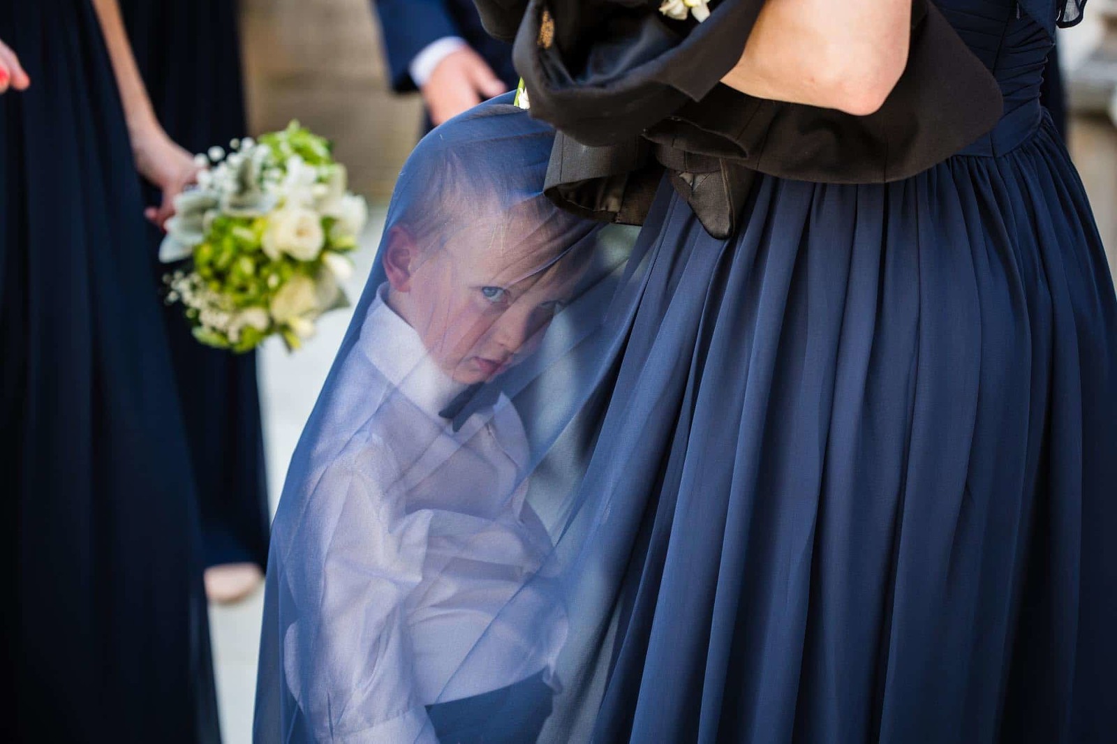 disgruntled ring bearer hiding under bridesmaid's dress, giving bored look