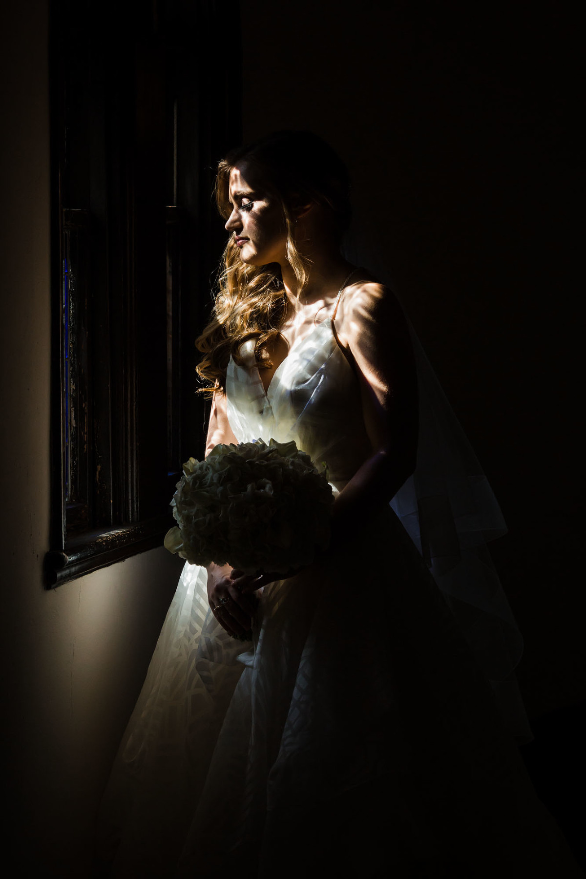 bride standing at the window, in spotty light. dramatic high contrast profile photo