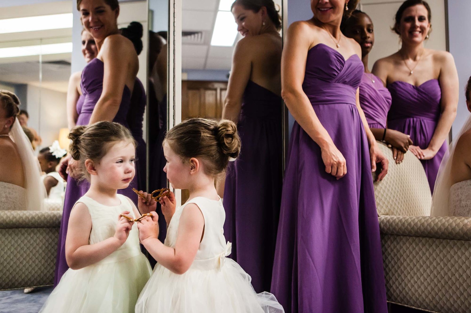 cute flower girl looking at herself in the mirror, with a pretzel, in the bridal suite at heinz chapel, before the wedding ceremony begins