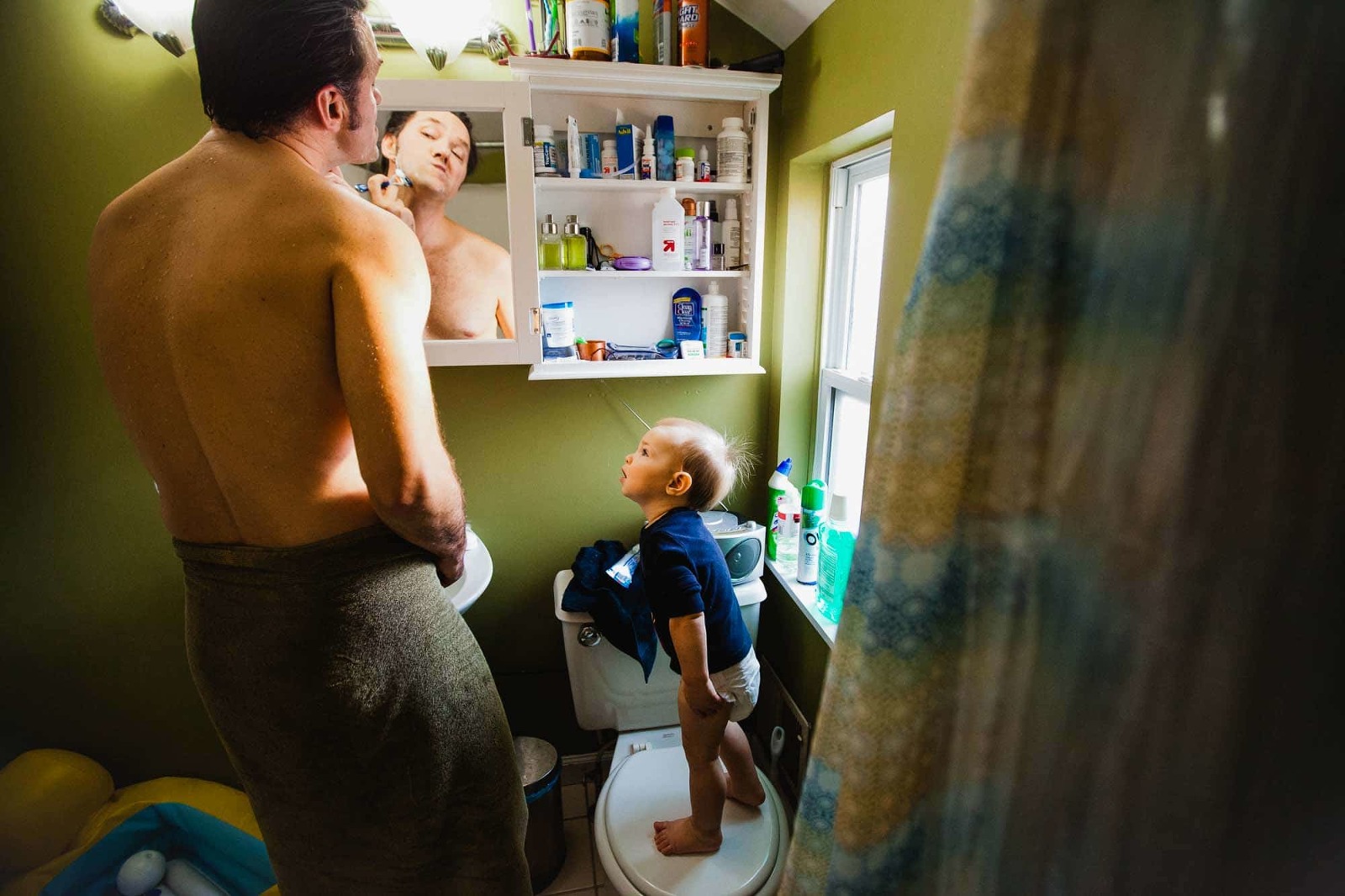 dad shaving shirtless in the bathroom with his toddler son standing on the toilet watching intently