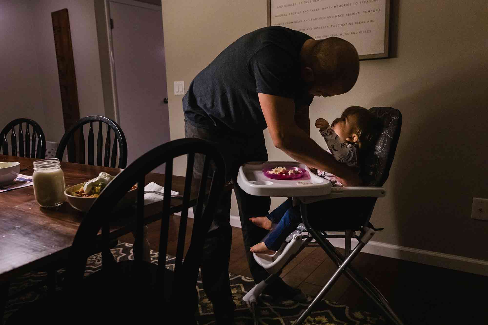 little girl in high chair reaching up for her dad as he is trying to get her out of the seat