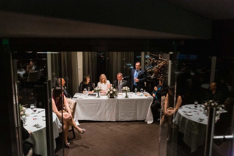 wedding reception toasts from best man, photographed from afar through doorway, with guests seated and laughing