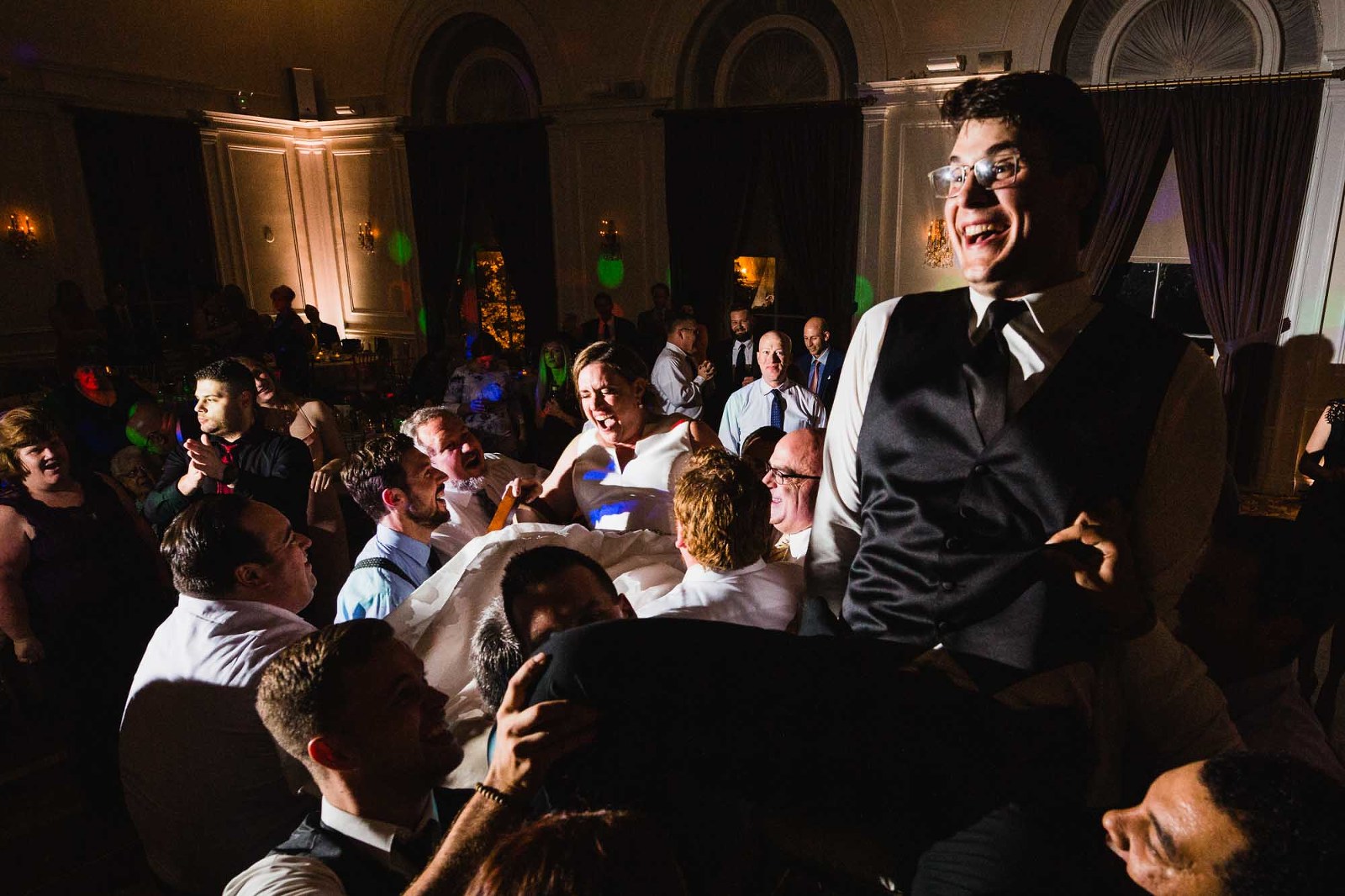 groom lifted up on chair during wedding reception, surrounded by cheering guests