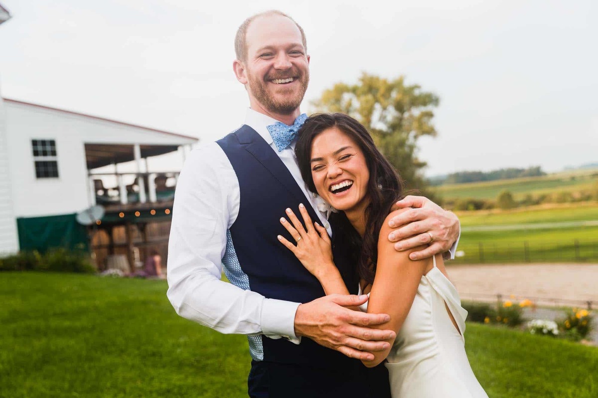 smiling groom hugs bride into his chest and they laugh together, surrounded by farm