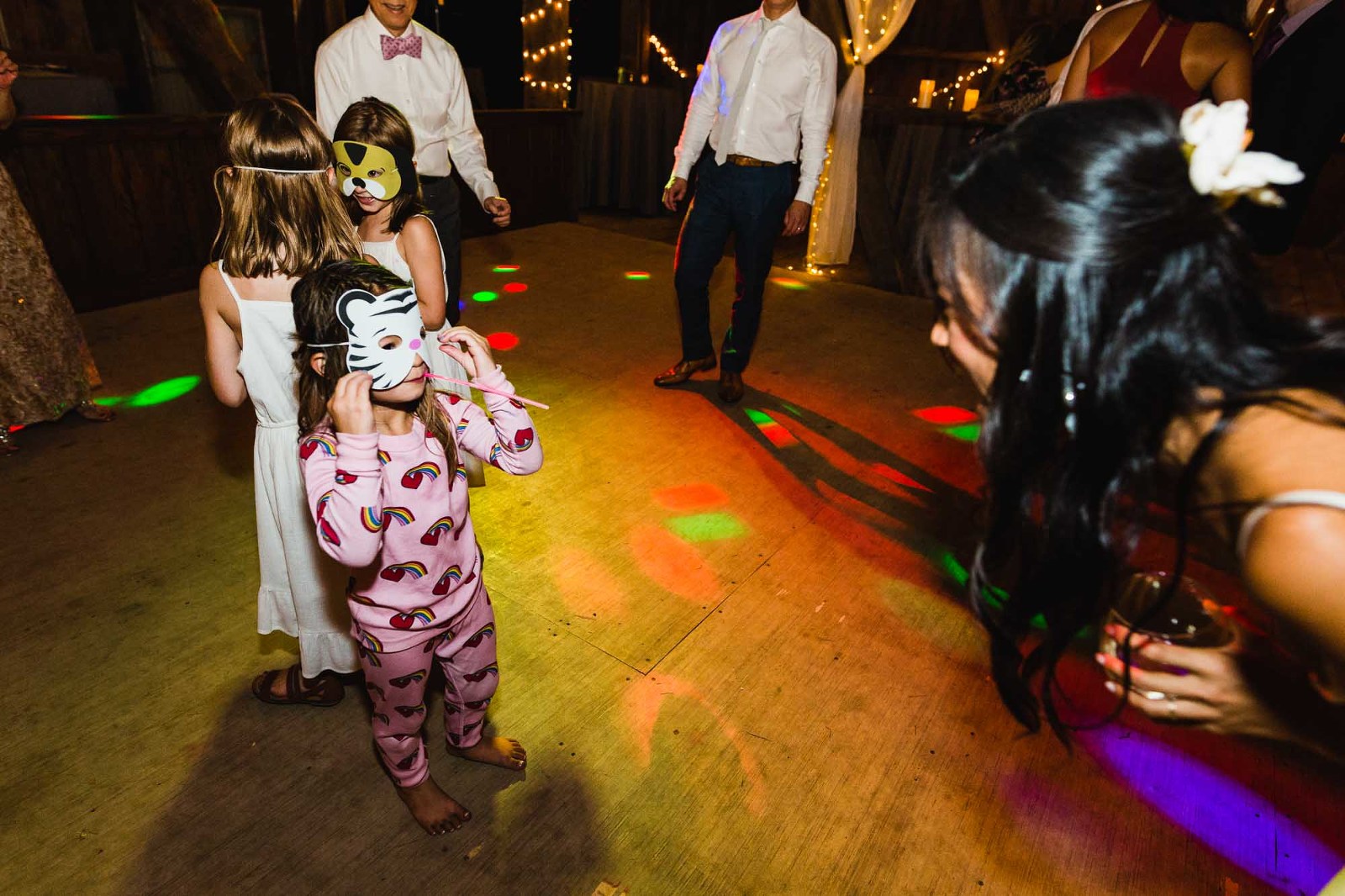 little girl in pajamas wears mask on wedding dance floor with lights flashing, looks up at bride