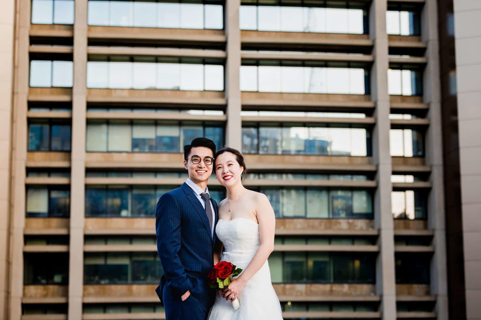 bride and groom pose for the camera in a parking lot in oakland, in front of a tall building on pitt campus