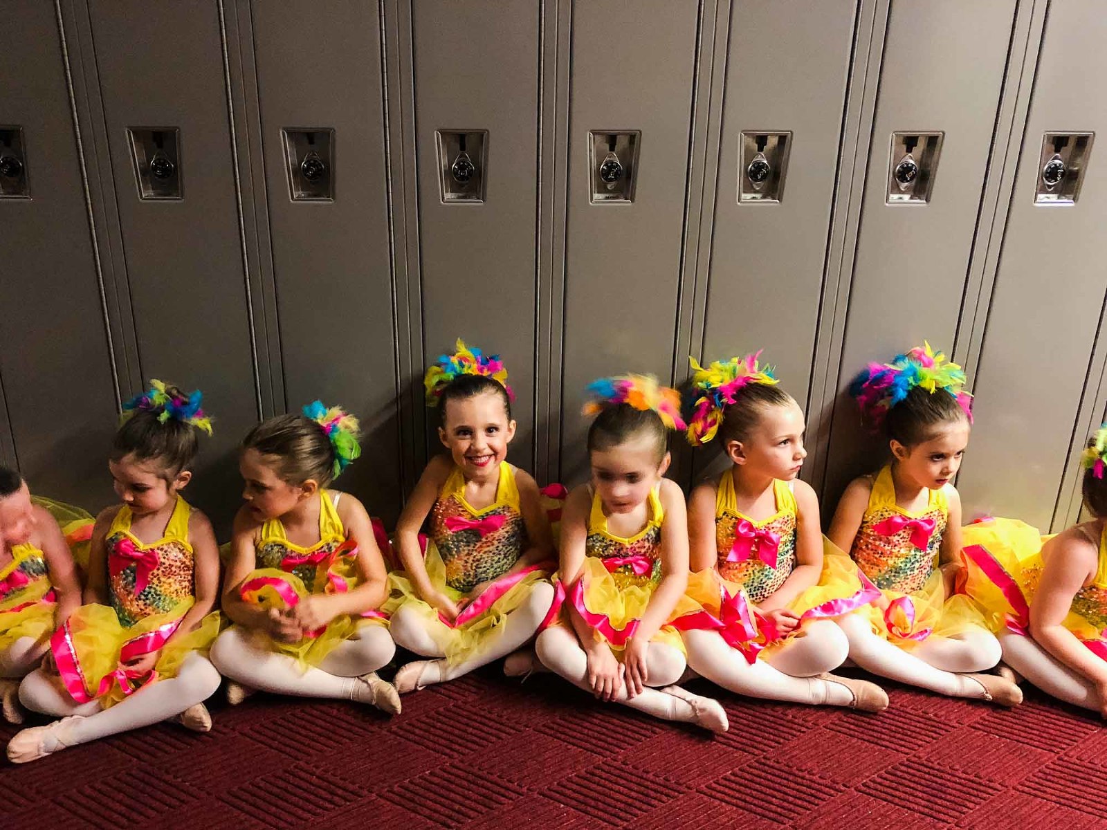 ballerinas lined up ready to go on the stage, in yellow and pink tutus, seated in front of a row of school lockers