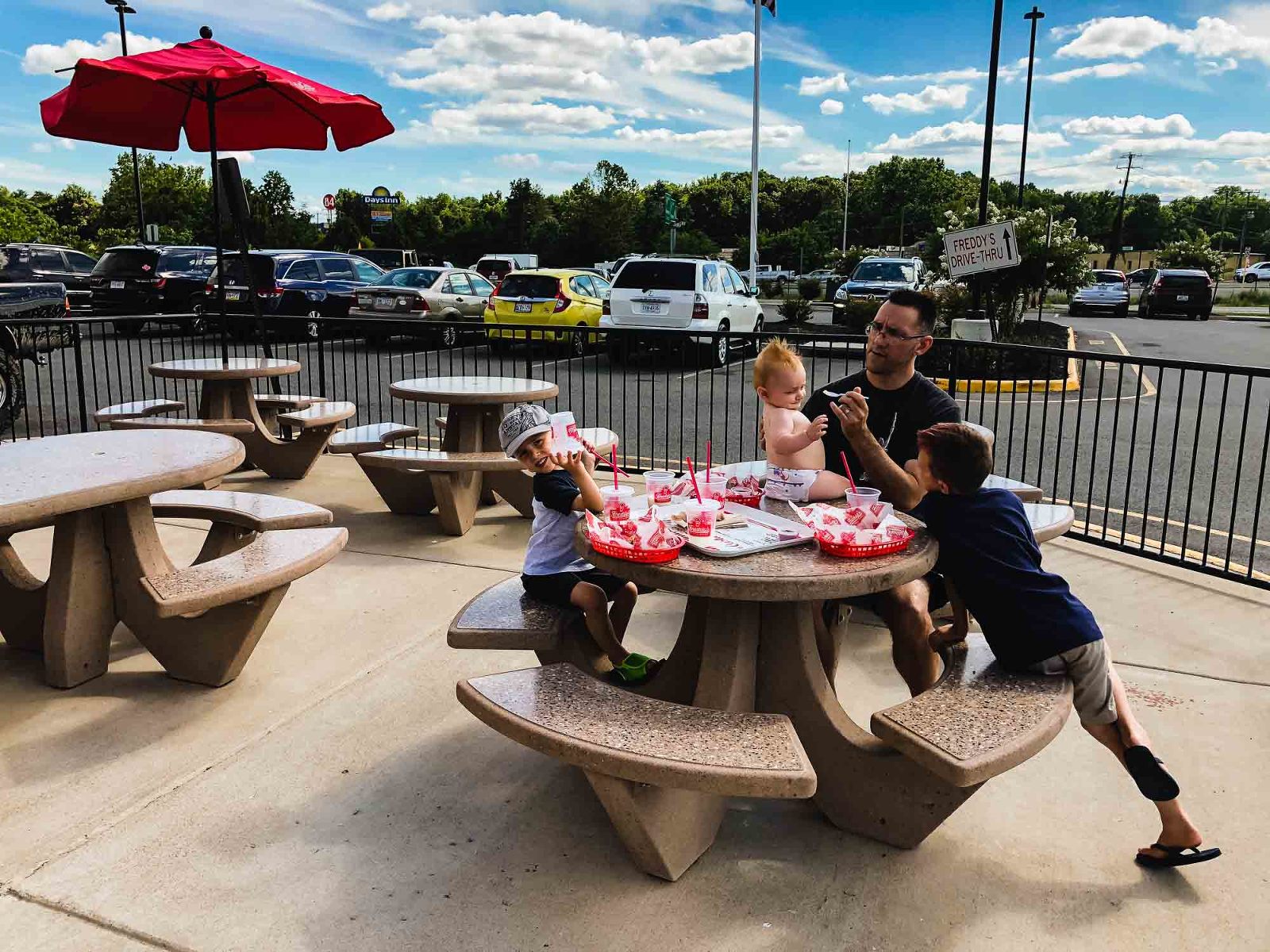 dad and three kids sit at a concrete picnic table outside a fast food restaurant, surrounded by parking lot, feeding a baby sitting on the table
