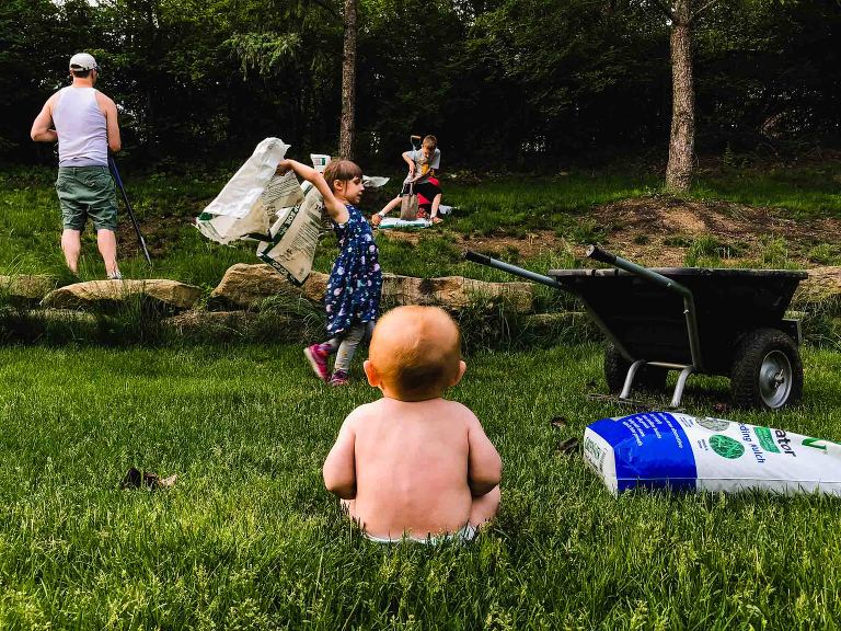 naked baby sits in grass watching family do yardwork in the yard beyond