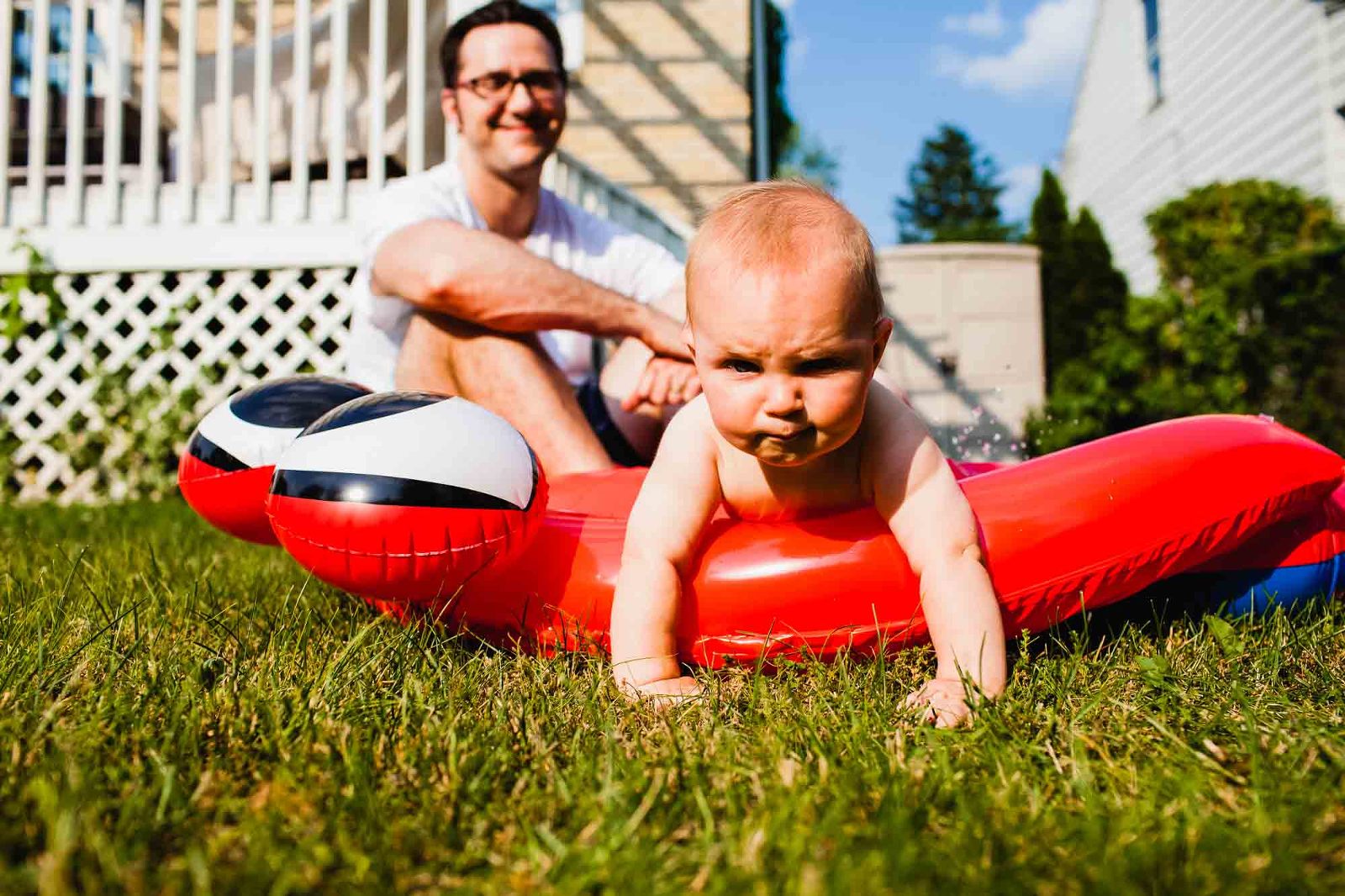 toddler climbs out of red backyard blow up pool, with disgruntled expression, and dad laughing behind him