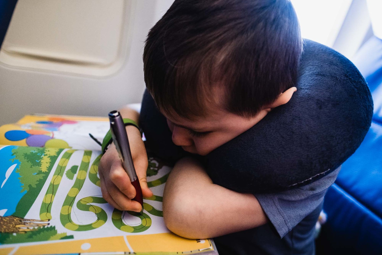 little boy on airplane wears sleeping pillow around his neck and does puzzle book