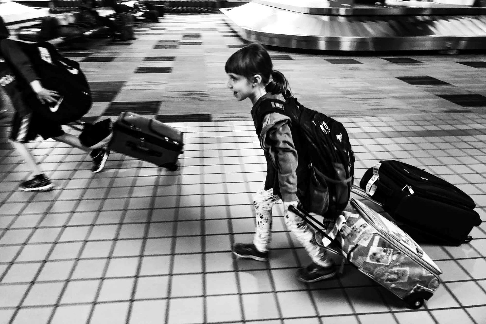 black and white photo of little girl walking through the airport carrying two rolling suitcases and a backpack