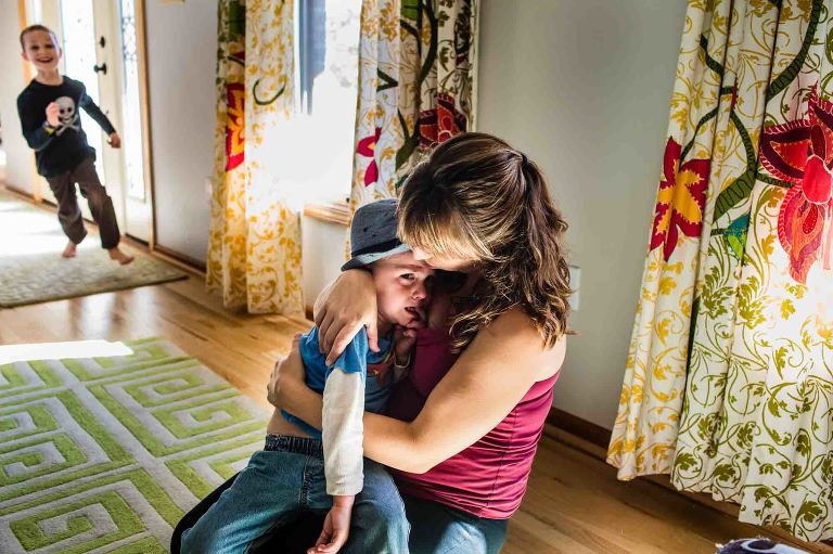 mom cradling crying boy on the floor of a middle class home while brother runs laughing through the room
