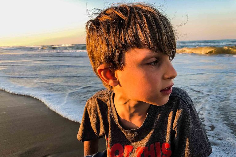 closeup of boy on the beach, looking away from the sun toward the water, medium length hair flying wildly