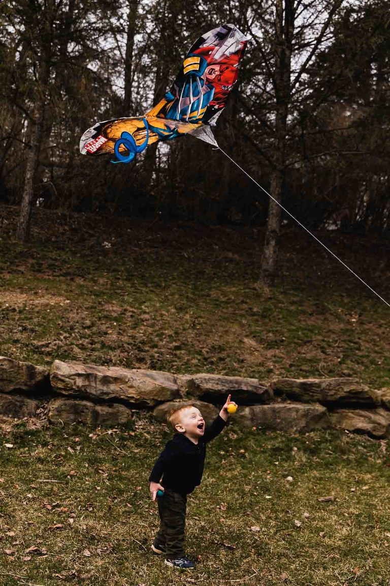 vertical color photo of toddler laughing and pointing up at a kite flying above him, in a mccandless backyard