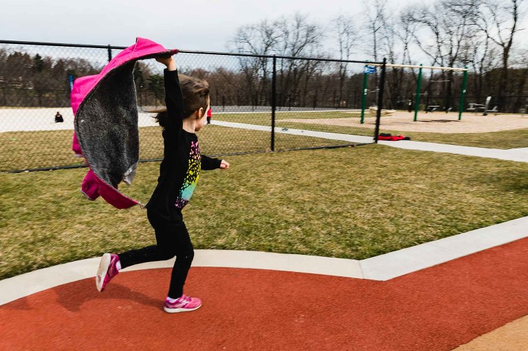 little girl with dark hair, black long sleeve clothes, and pink shoes runs through a playground with her arm in the air clutching her coat flailing in the wind. photograph taken with movement
