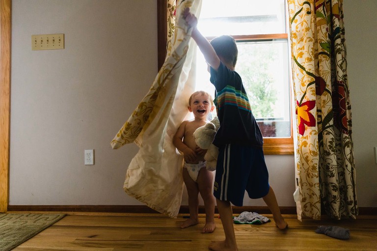 toddler in diaper hides behind curtain as boy pulls curtain away to find him. both boys laughing