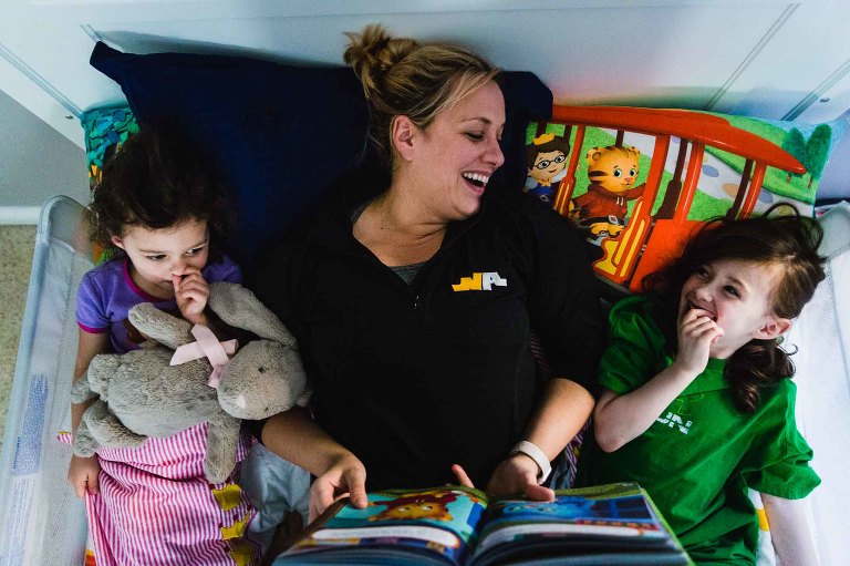 mom lies in bed with two young daughters, reading bedtime stories. laughing with one and the other picks her nose