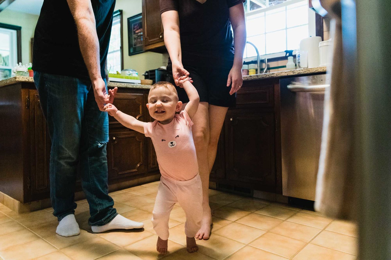 baby learning to walk holds onto mom and dad's hands as she walks through her kitchen