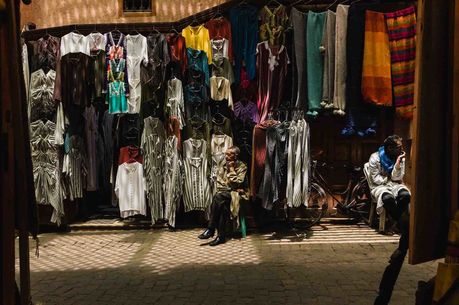 moroccan street vendors sitting in spotty light surrounded by colorful clothes on a narrow street