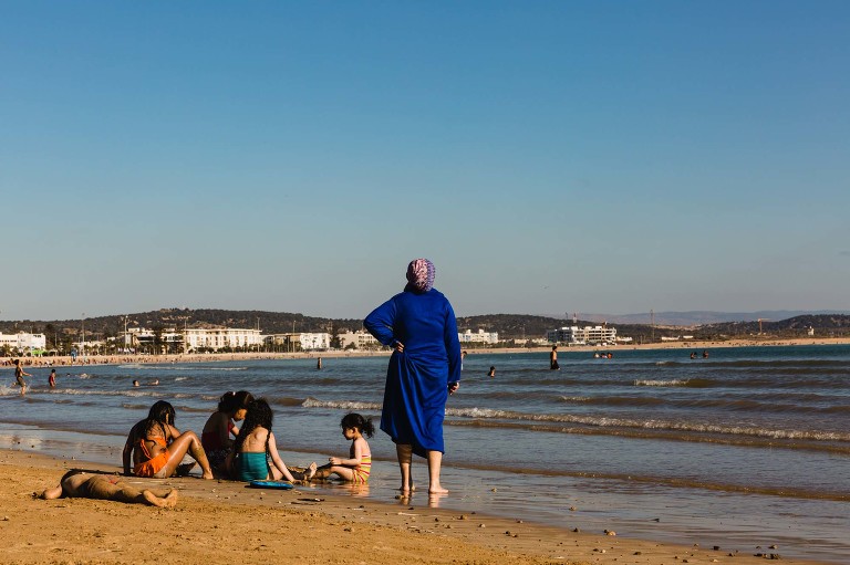 mom in robe stands on beach with hand on hip, kids playing beside her, gazing out over the beach water