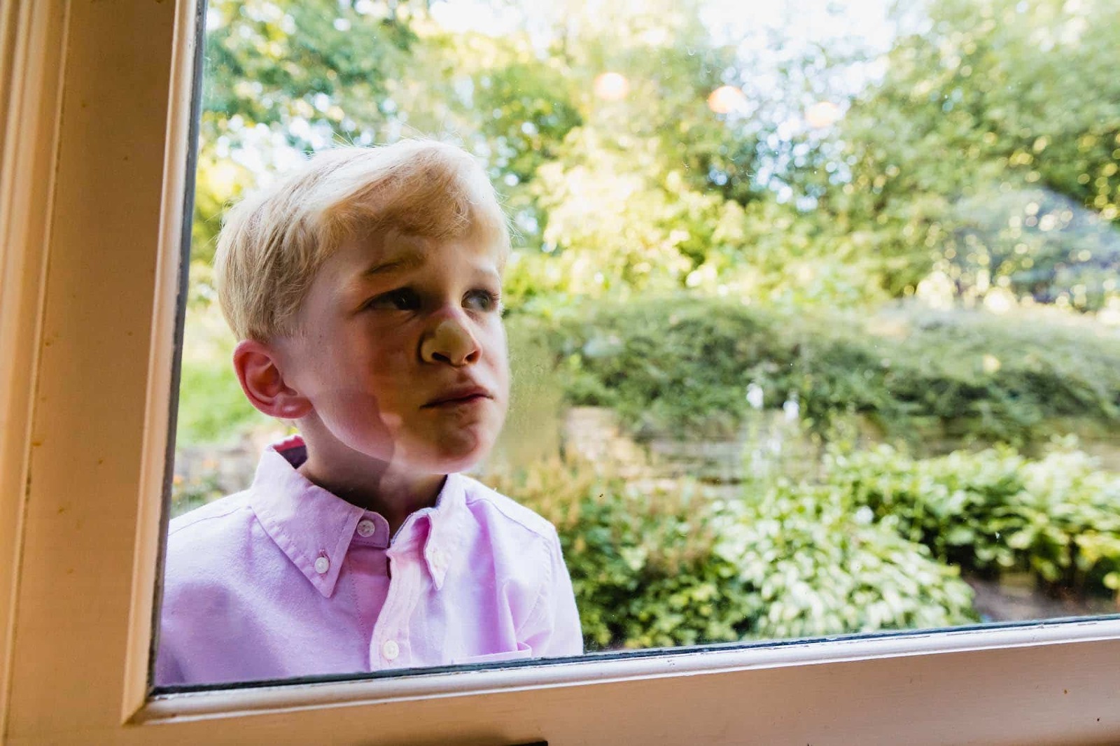 young boy in pink button-down shirt presses his face up against a window, in front of green backyard. 