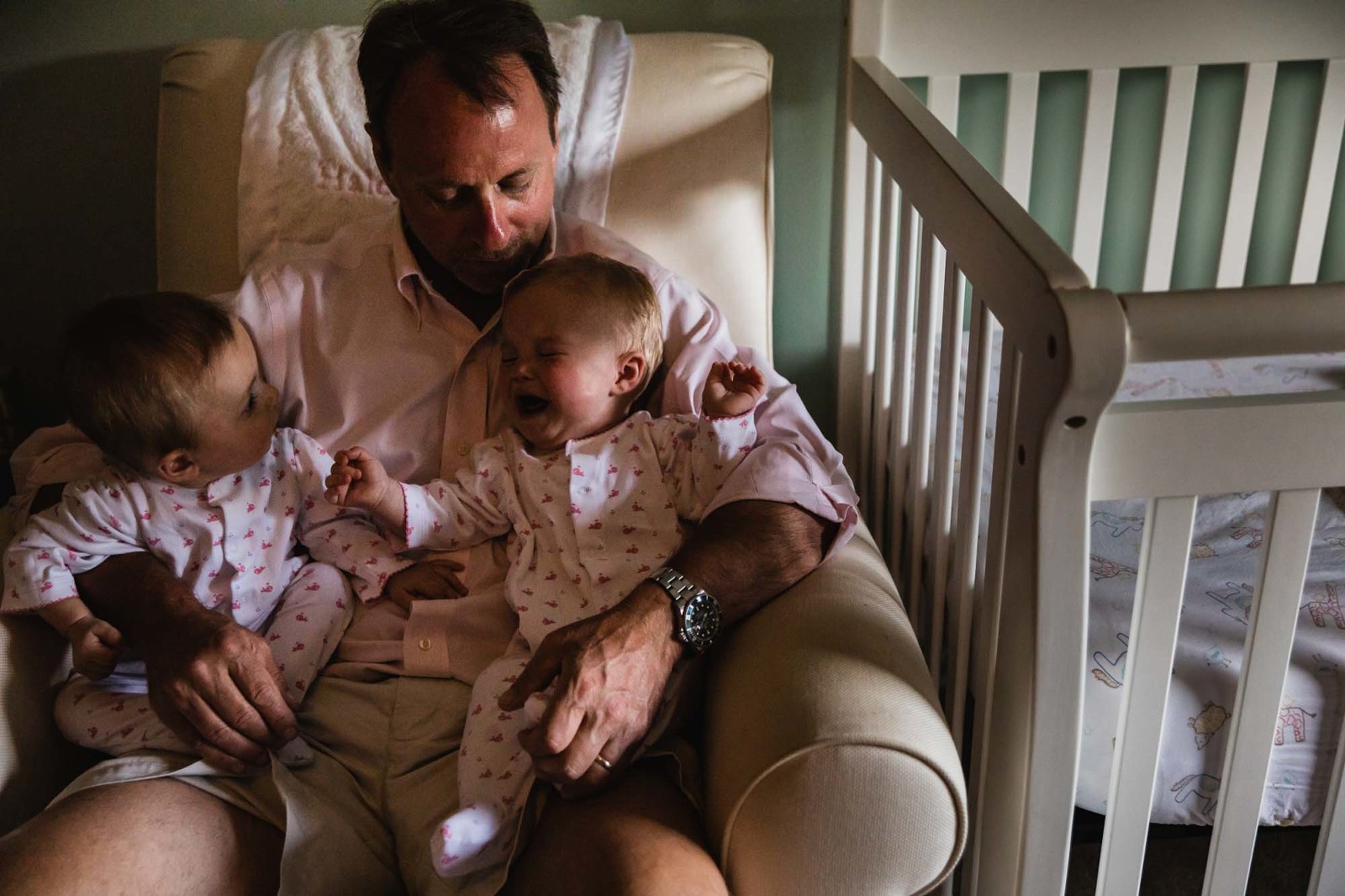 dad holds his twin daughters on his lap. one little girl cries and the other looks on confused.