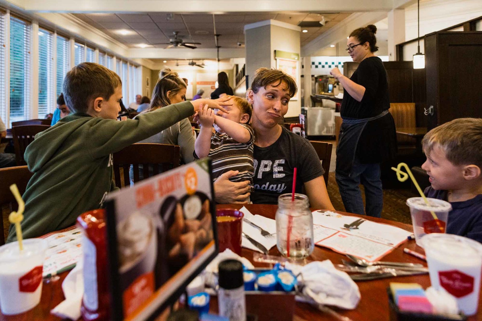 family eating in restaurant, mom holding baby and talking to boy while another boy plays with toddler