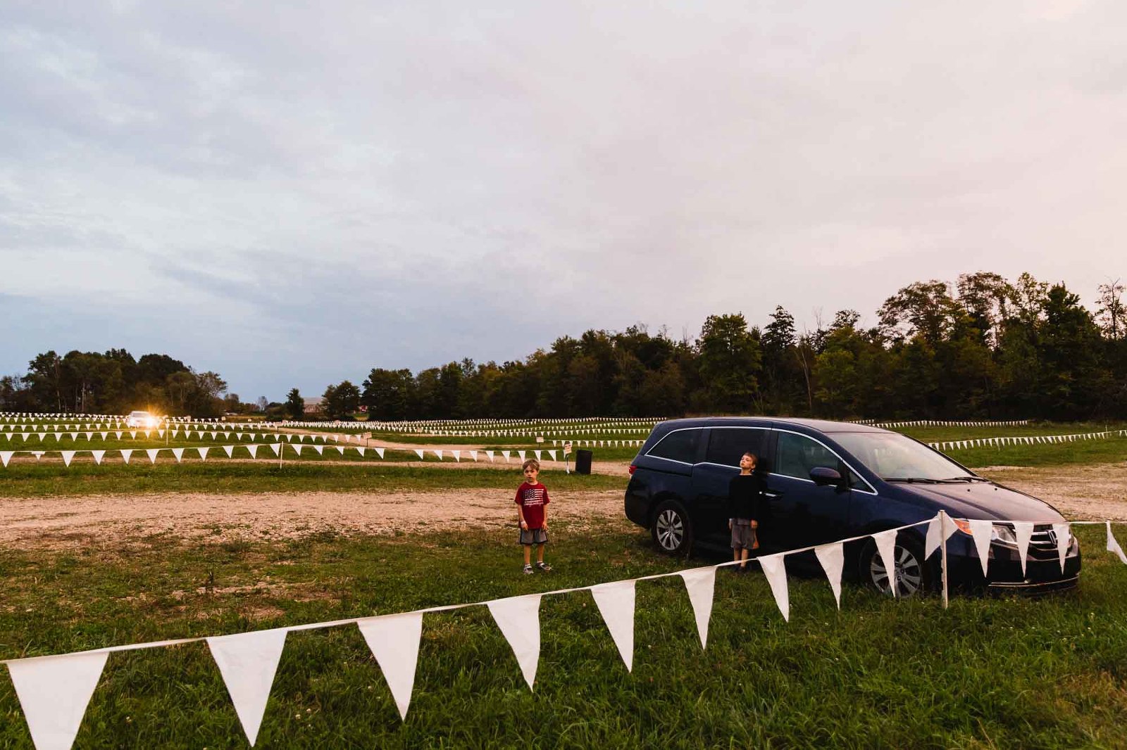 lone car sits in a giant makeshift field parking lot at sunset, with two boys lounging
