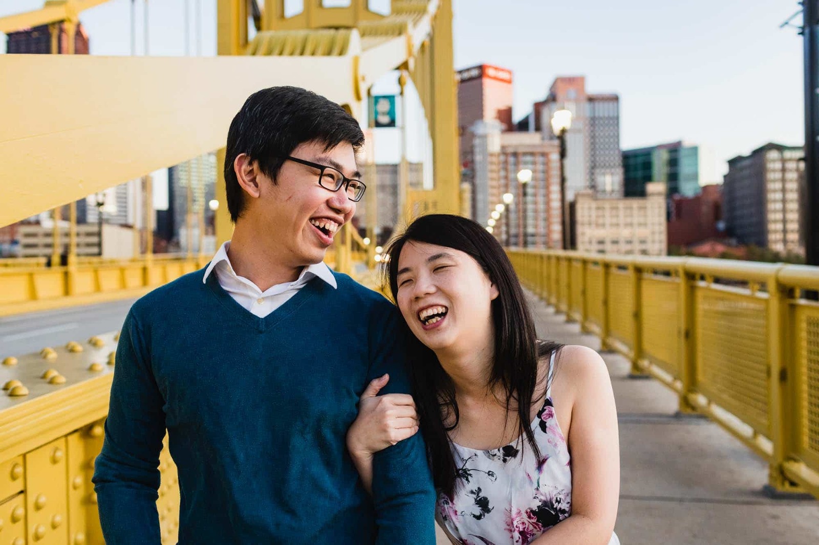 asian couple laugh together joyfully, framed by yellow bridge and city of pittsburgh