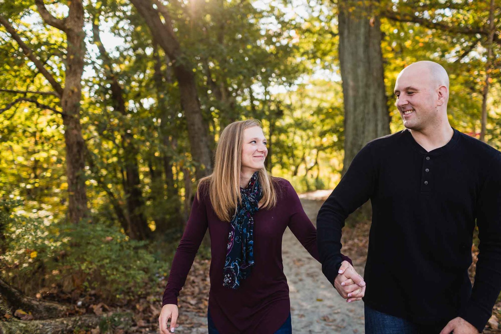 woman and man holding hands and walking a path in the woods, smiling at each other