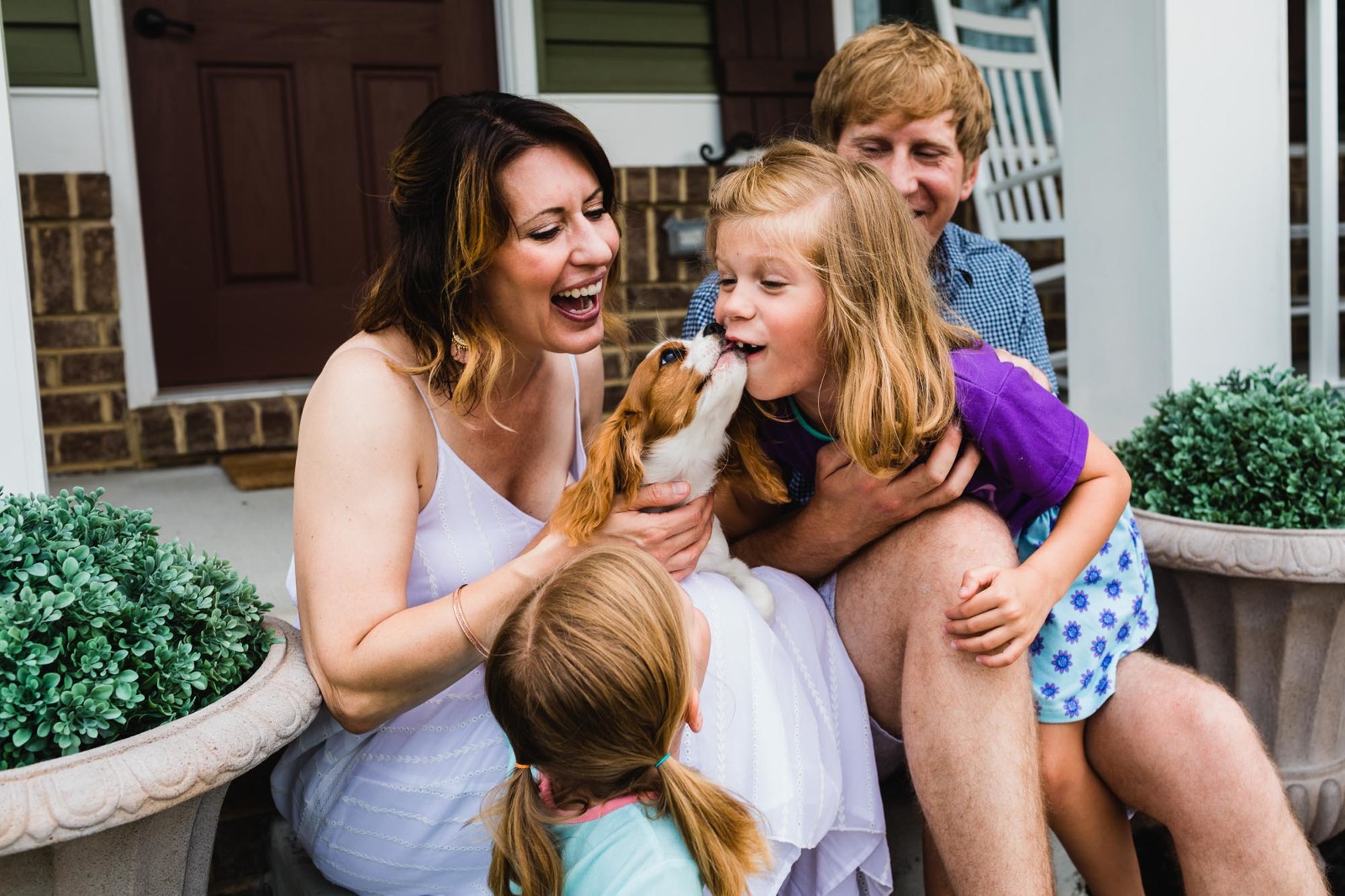 dog licking little girl's face, sitting amongst her family members on their front porch, with everybody laughing
