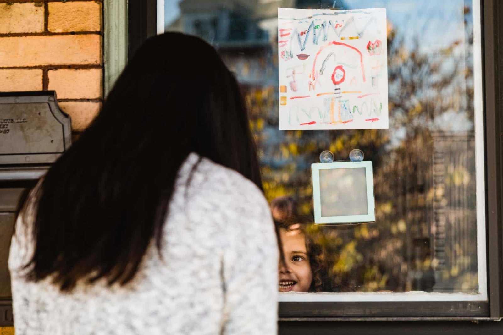 little girl smiles proudly out the window of her home, with her art hanging on the window above her head