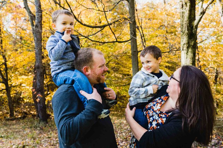 toddler on dad's shoulders, boy in mom's arms, all laughing together and looking at each other, in front of fall foliage