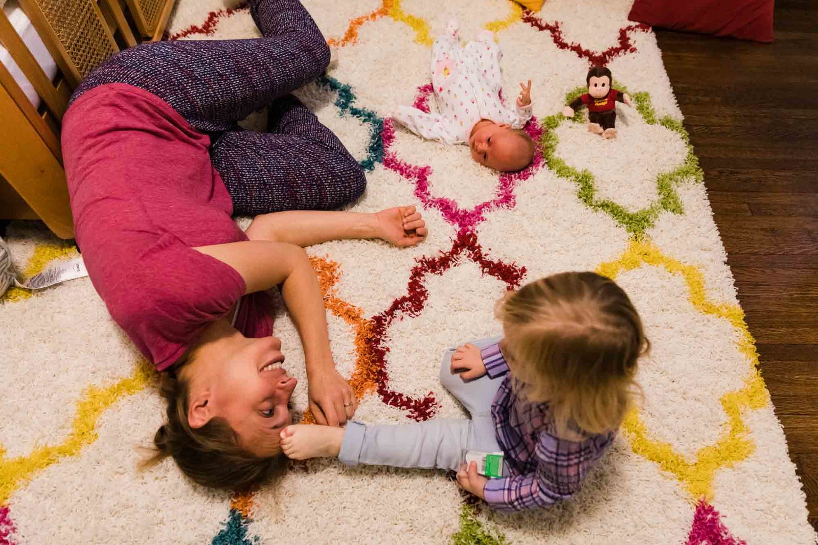 little girl pushes her foot into her mom's head as mom lies on the carpet. baby lies nearby. photo taken from above. 