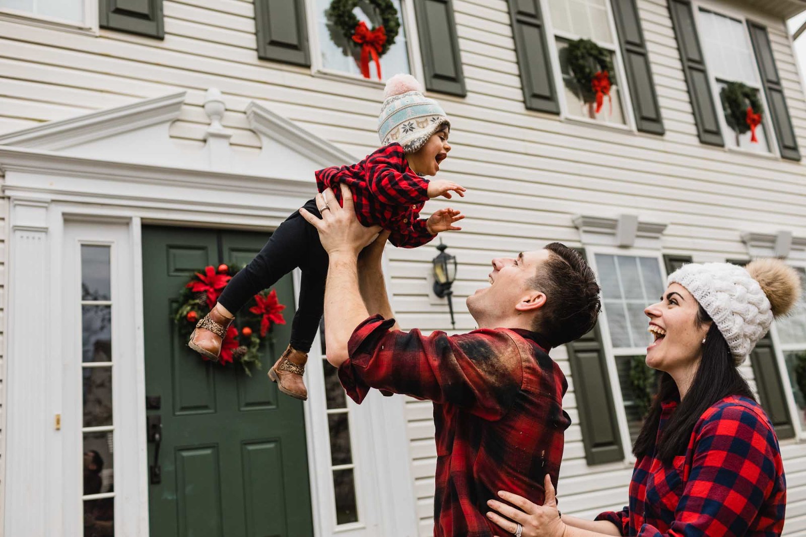 dad throws baby into the air, and mom holds dad from behind also laughing. house in the background. 