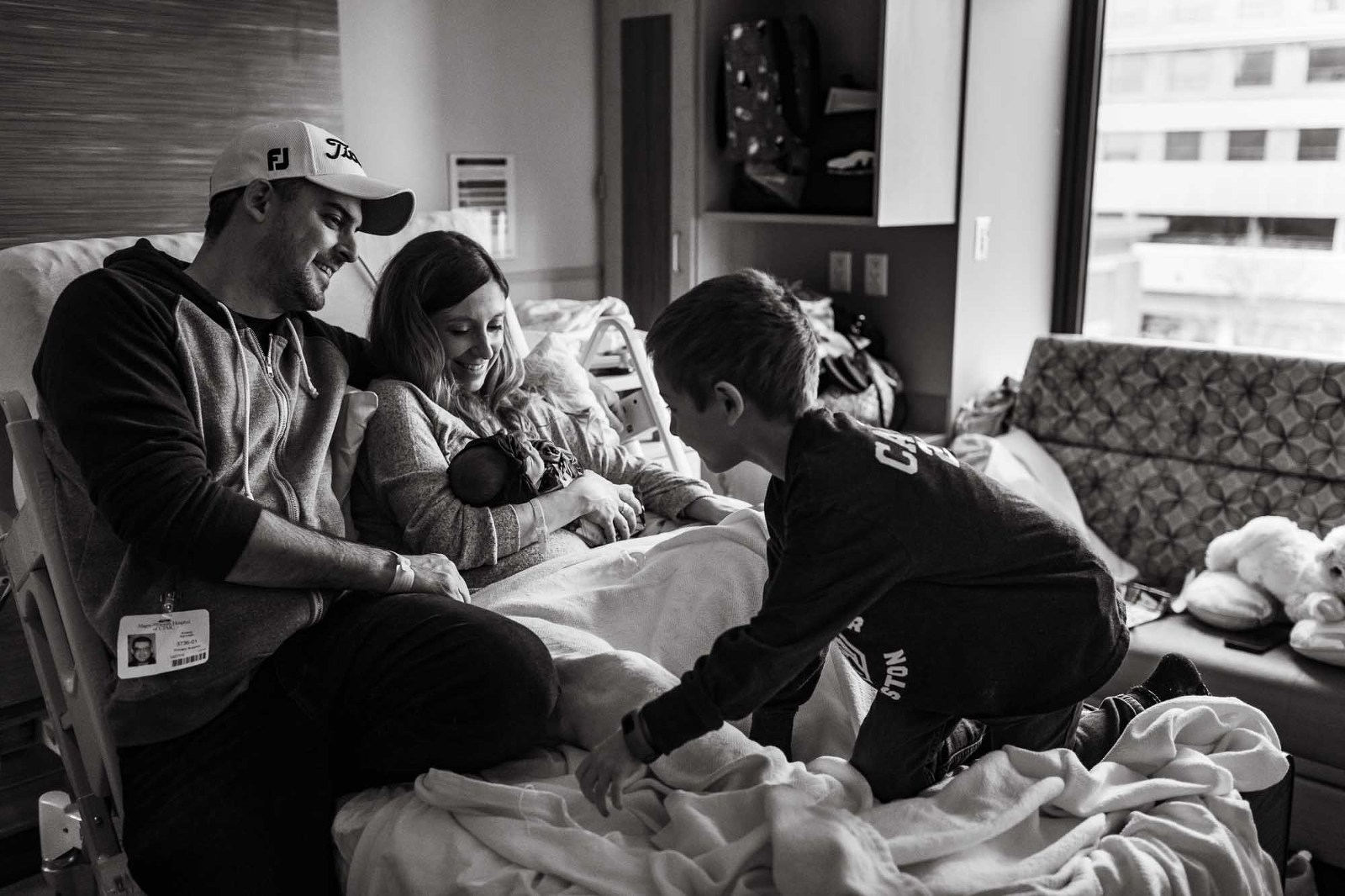 black and white photo of boy climbing up onto hospital bed to join his parents and new baby sister