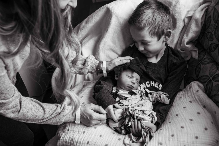 big brother holds baby sister, smiling, while mom reaches in