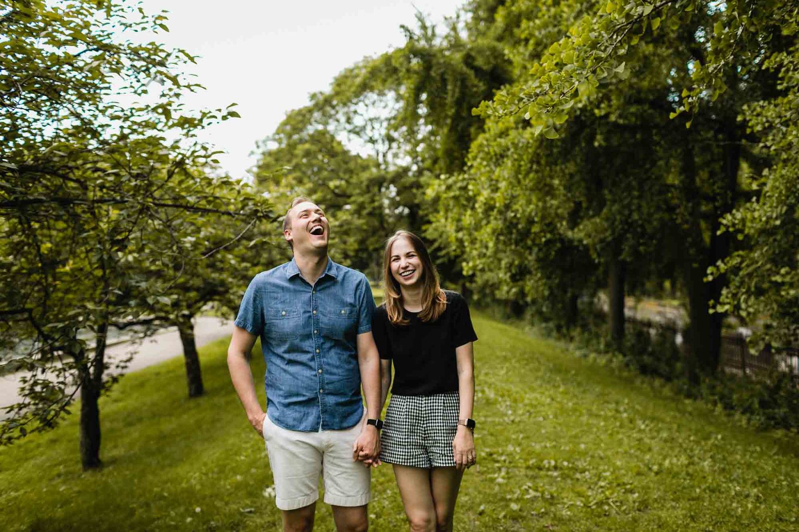 man and woman stand next to each other holding hands, laughing, framed by some trees in the park. 