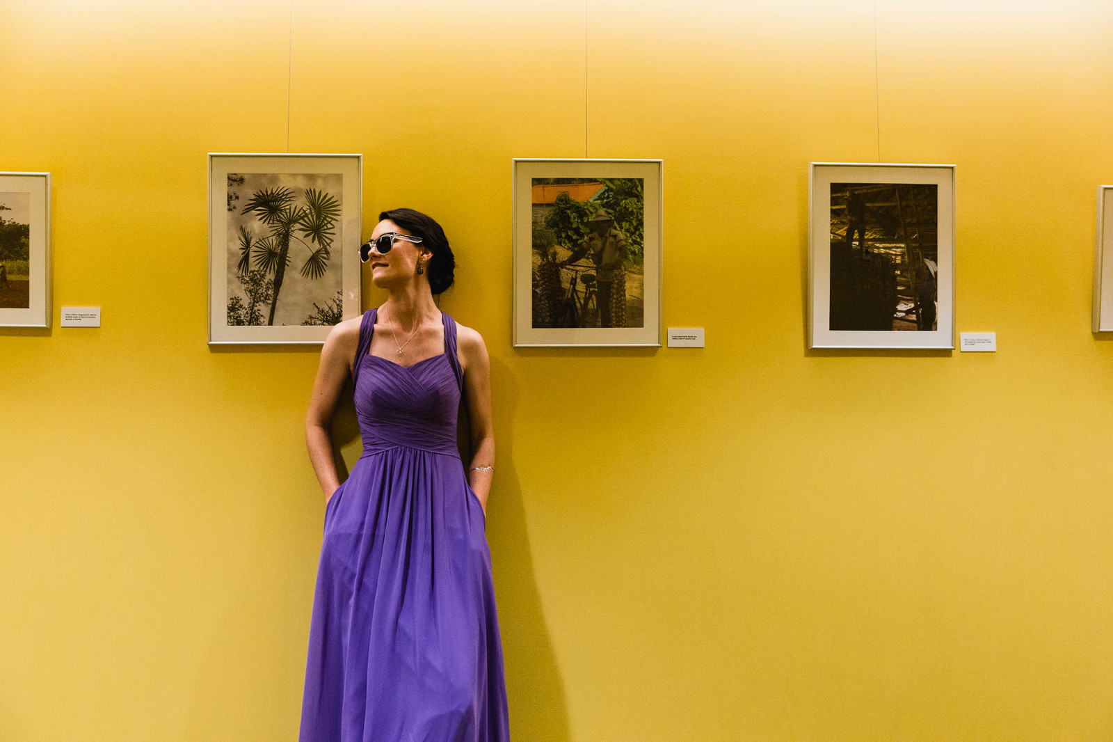 Bridesmaid wears sunglasses and leans against a wall covered in portraits. 