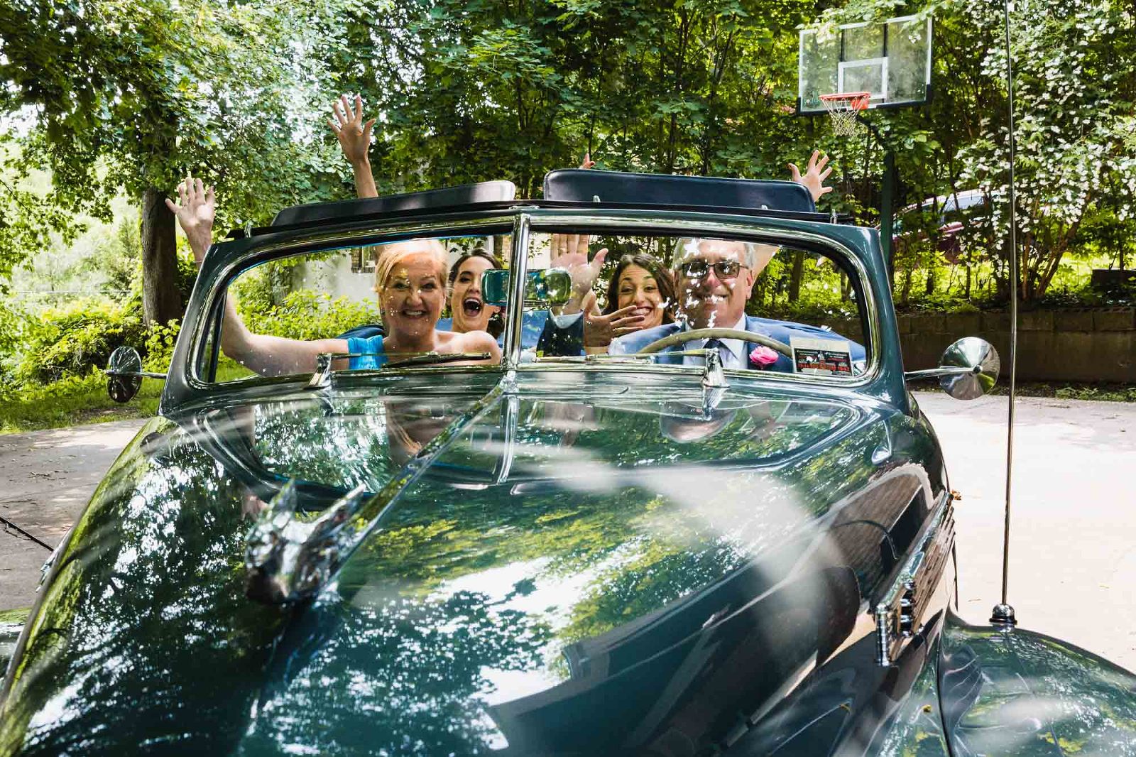 family cheering for the camera, seated in an antique jaguar car, in front of a basketball hoop
