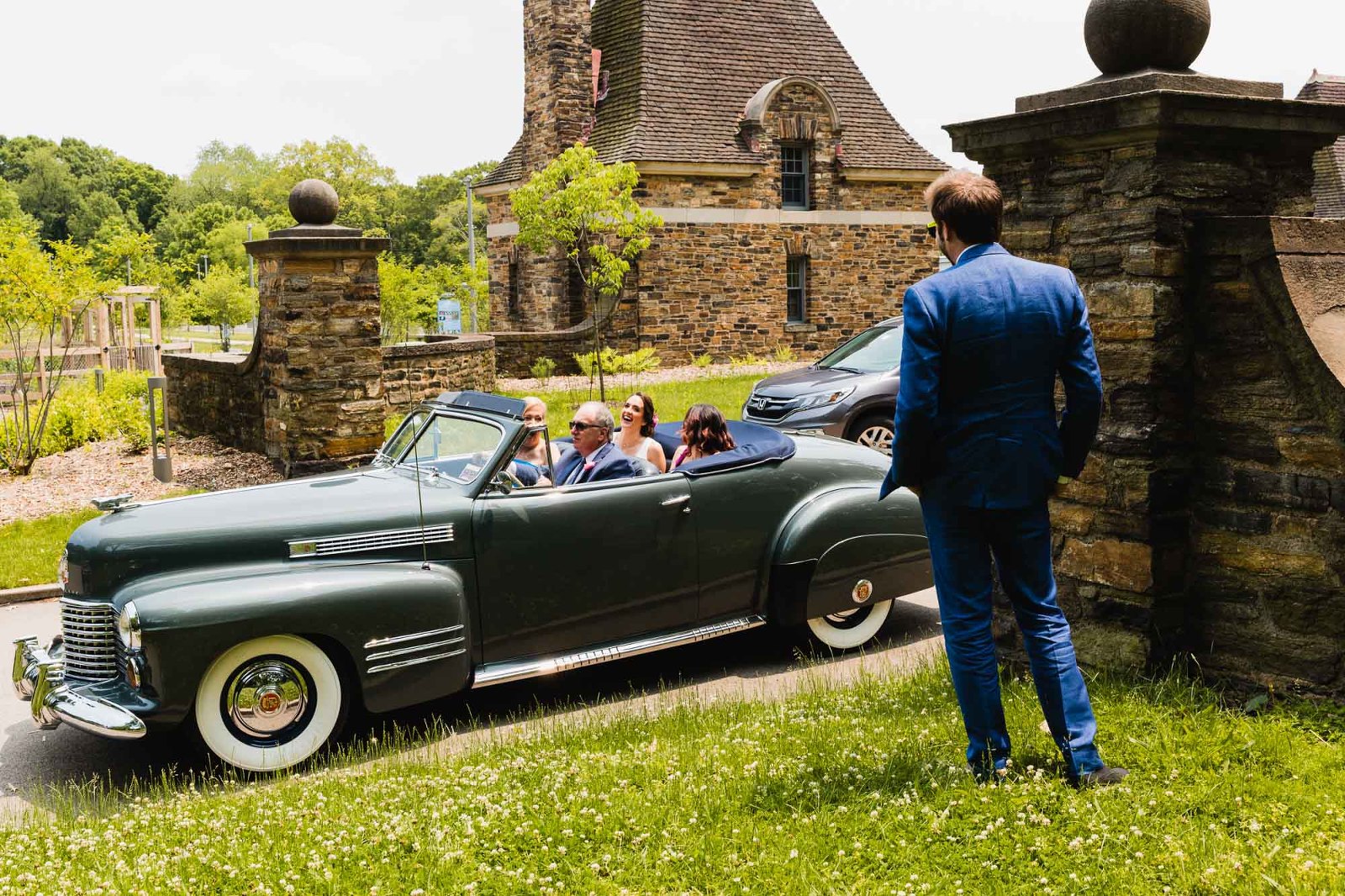 bride rides in an antique jaguar car with her parents, waving to her groom standing nearby