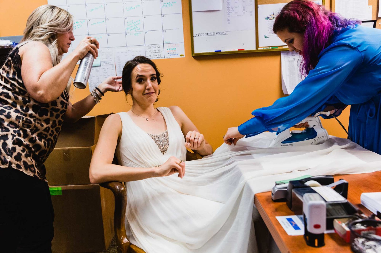 bride before her wedding ceremony, fully dressed, with friend ironing her dress on an office desk, and another friend fixing her hair