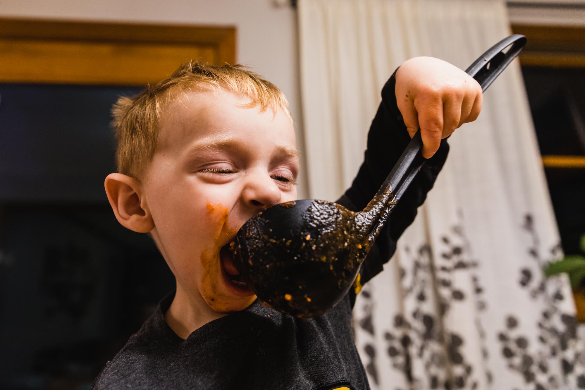 toddler holds soup ladle up to his mouth for a drink, eyes squeezed shut and mouth wide open, face covered with red sauce