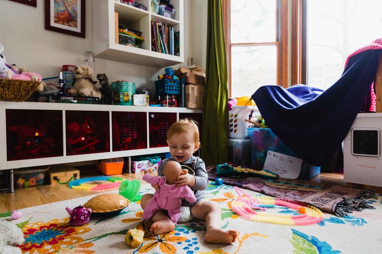 toddler plays with baby doll, sitting on the floor of a messy play room